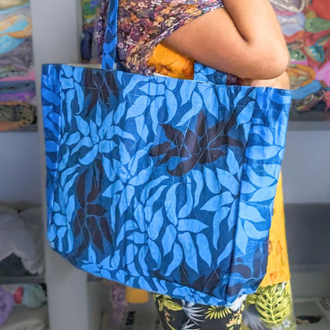 A large tote bag made from upcycled sari fabric, held by a person with a colorful garment in the background. The bag features a geometric pattern in shades of blue.