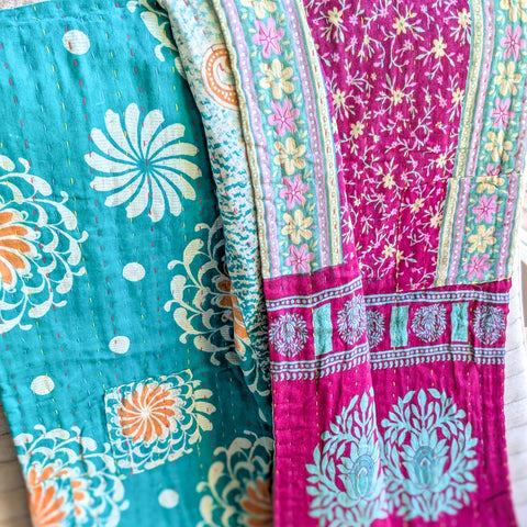 Two sides of one colourful patterned kantha quilt, one turquoise and one pink with floral designs, on a wooden surface.