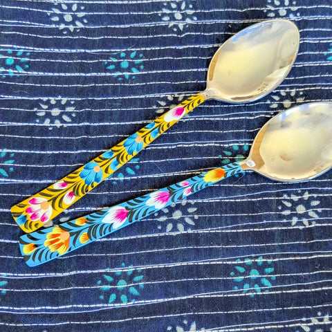 Two tablespoons with colourful painted handles on a blue patterned fabric background